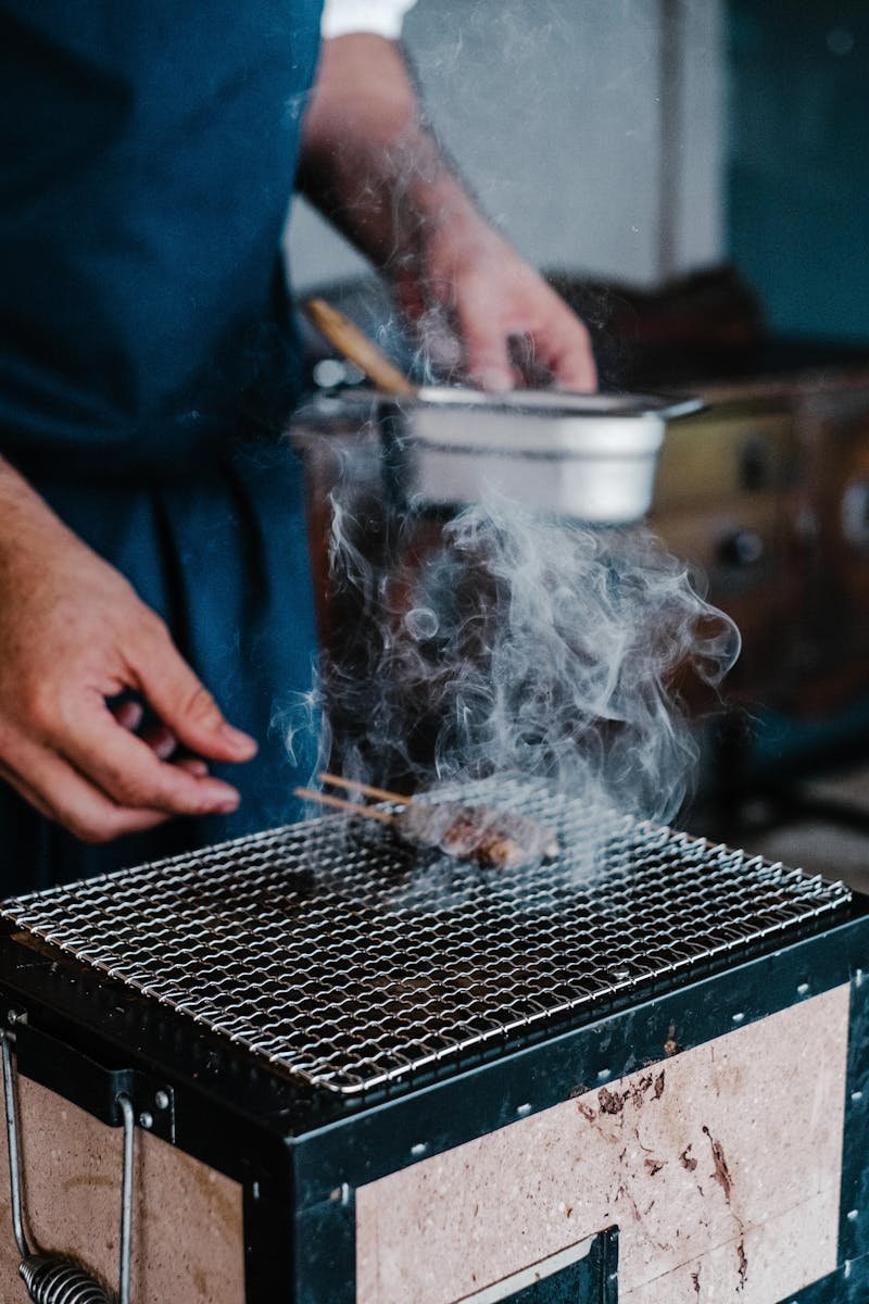 Close-up of grilling kebabs over a smoky barbecue, showcasing hands in action.